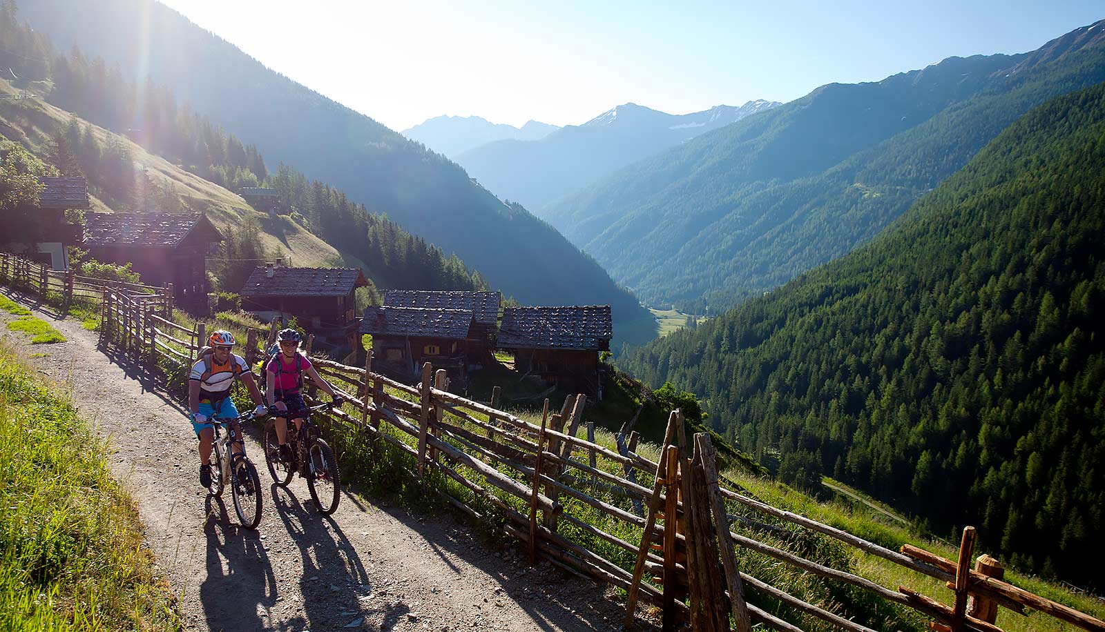 Zwei Mountain Biker auf einem Weg hinter Bauernhöfen zwischen den Bergen an einem Sonnentag