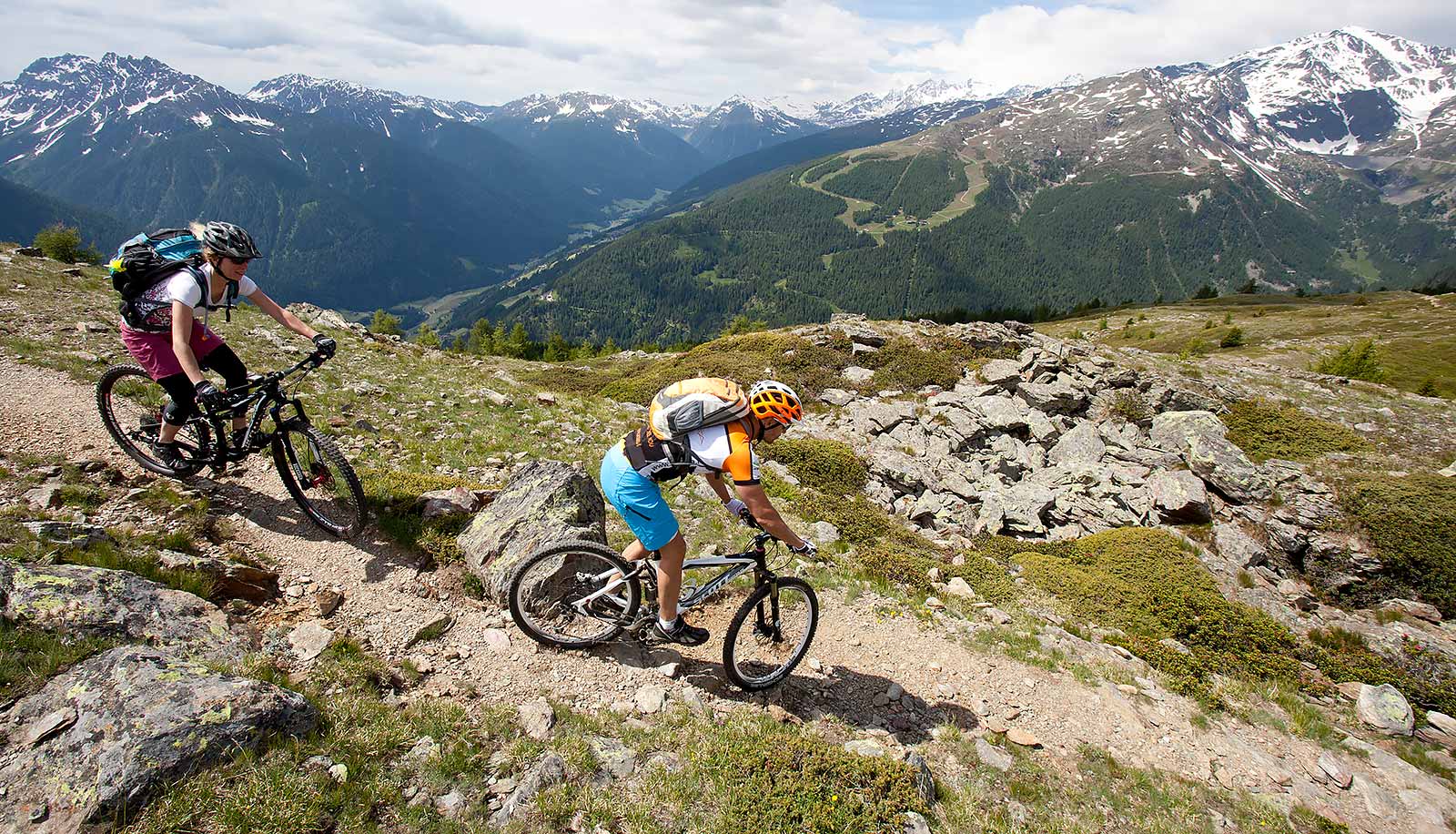 Zwei Mountain Biker auf einem steilen Bergweg inmitten der Felsen mit verschneiten Bergen im Hintergrund